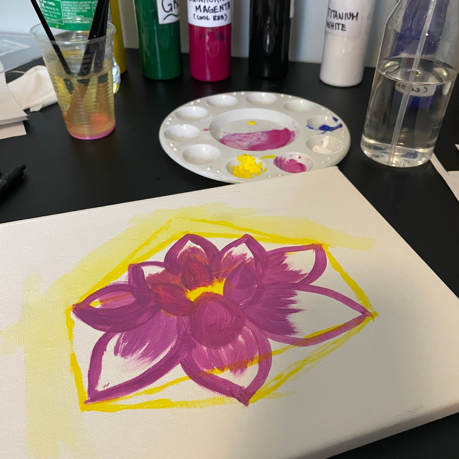 Art supplies including paint bottles, a palette, and a painting of a purple flower on a white surface.