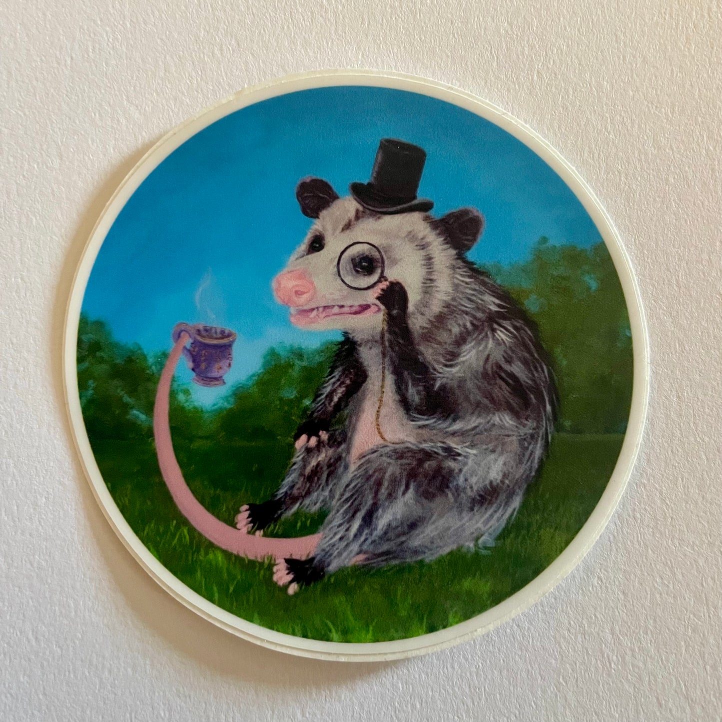 Sticker with an illustration of an opossum wearing a top hat and holding a tea cup on a grassy background.